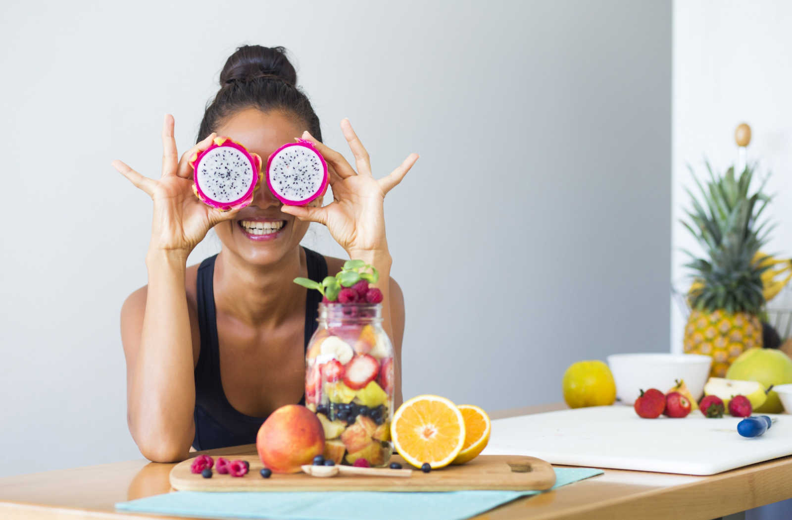 A smiling woman holds two two pieces of dragonfruit in front of her eyes, with other cut up fruits laid in front of her on a table.